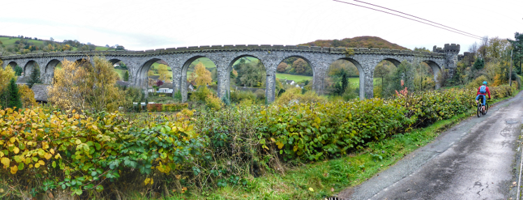A panoramic shot of a cyclist riding along the road with an impressive viaduct in the background
