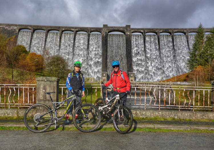 Two cyclists pose with their mountain bikes beneath a dam on the Trans Cambrian Way